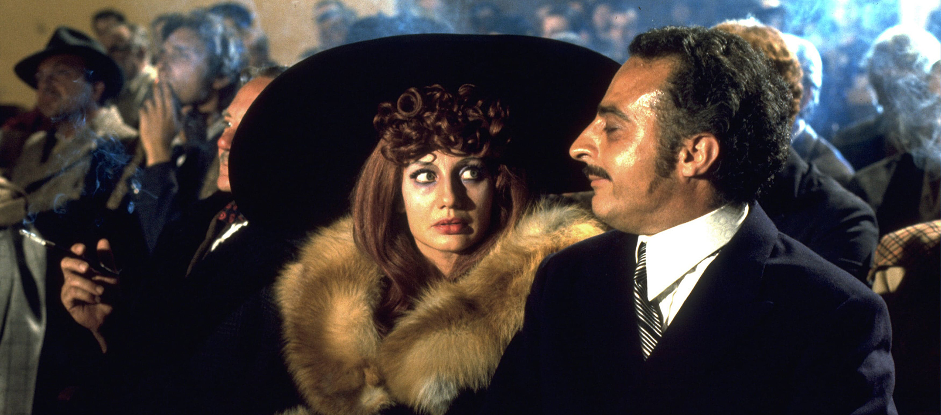 Still featuring Elvira (Cassandra Peterson) sitting next to a man in an audience. She has brownish red hair with curled bangs and is wearing a large black hat and fur coat.