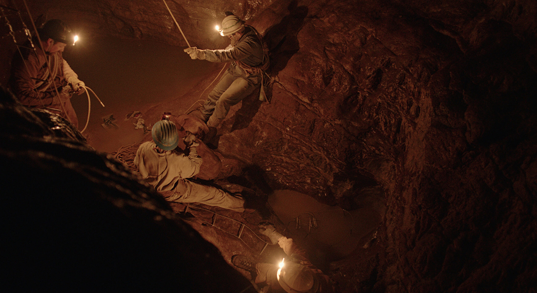 Four people in a rocky cave lit by the minor hats they’re wearing. They have rope tied around them as if to descend.