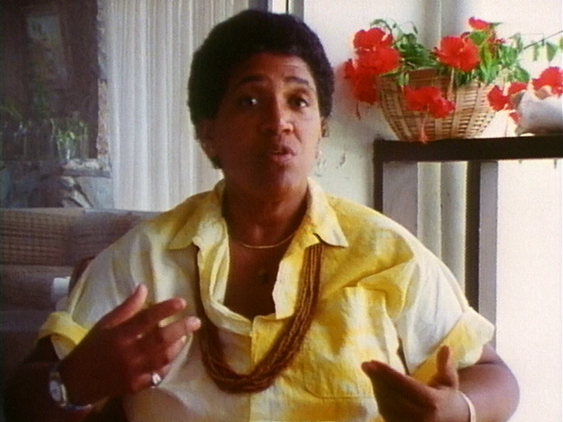 Audre Lorde is seated and wears a yellow shirt, unbuttoned at the neck. Behind them is a vase of red flowers on a table.