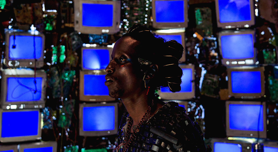 Profile view of Bertrand Ninteretse in Neptune Frost. They have red and blue paint around their eye, golden wire wrapped in their hair, and multicolored wire jewelry on their ears and neck. Behind them is a wall filled with rows of blue computer screens.