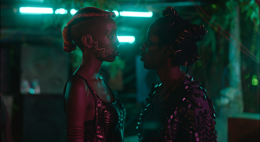 Cheryl Isheja (left) and Bertrand Ninteretse (right) facing each other and looking into each other’s eyes in Neptune Frost. The side of Isheja’s head is shaved, and they have reddish-orange braids running from the top to the back of their head. They are wearing a wire earpiece, wire arm cuff, and metallic pink tank top. Ninteretse has wire wrapped in their hair and is wearing a purple shirt with matching-colored squares on it.