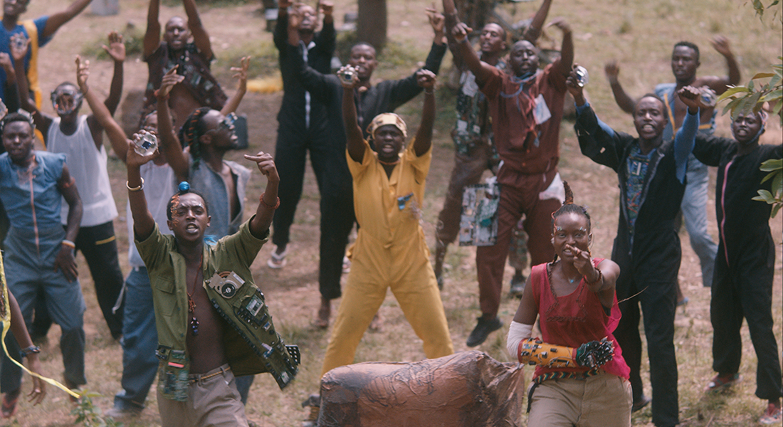 Group of Black people in a brown grassy field, looking into the camera as if in protest. They have their hands in the air—some with their middle fingers up, others with their hands in fists, and a few holding small devices.