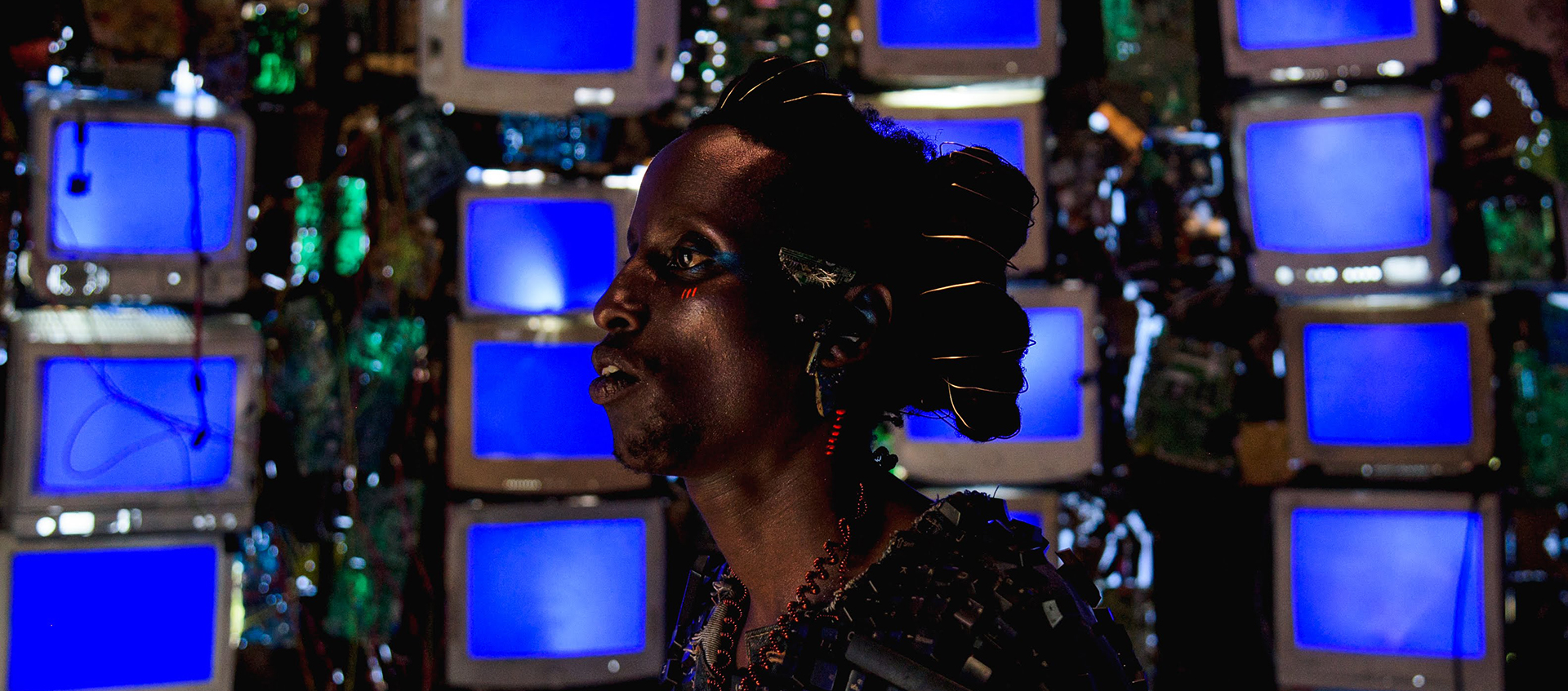 Profile view of Bertrand Ninteretse in Neptune Frost. They have red and blue paint around their eye, golden wire wrapped in their hair, and multicolored wire jewelry on their ears and neck. Behind them is a wall filled with rows of blue computer screens.