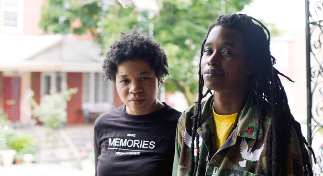 Rasheedah Phillips (left) in a black t-shirt and Camae Ayewa (right) in a camo jacket with roses and white doves on it. There is a red house and trees blurred in the background.