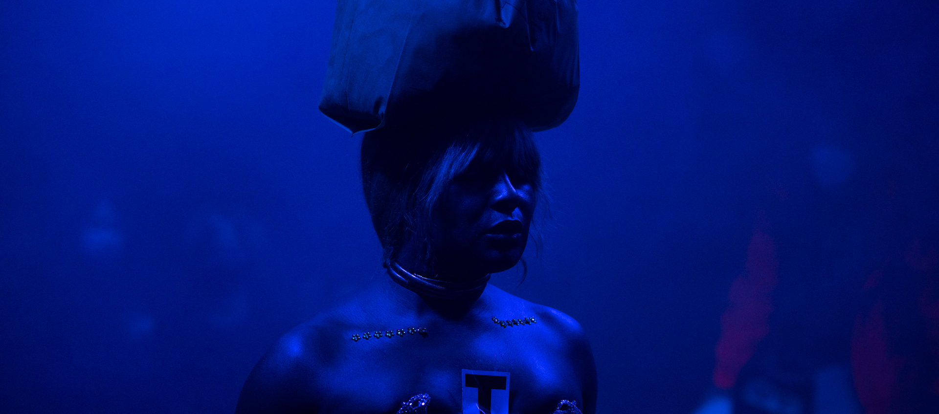 Artist Keioui Keijaun Thomas performing Comme Hell or High Femmes at Performance Space New York. Her shoulders are bare, and there are two separate lines of floral-shaped rhinestones on her chest. On top of her head, she wears a plastic bag with the handles tied. The entire room is bathed in blue light.
