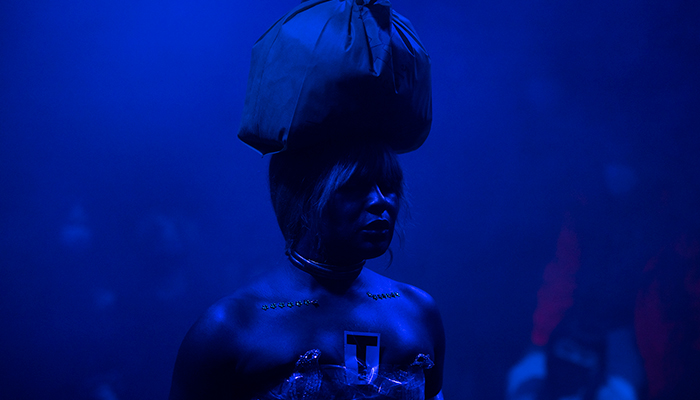Artist Keioui Keijaun Thomas performing Comme Hell or High Femmes at Performance Space New York. Her shoulders are bare, and there are two separate lines of floral-shaped rhinestones on her chest. On top of her head, she wears a plastic bag with the handles tied. The entire room is bathed in blue light.