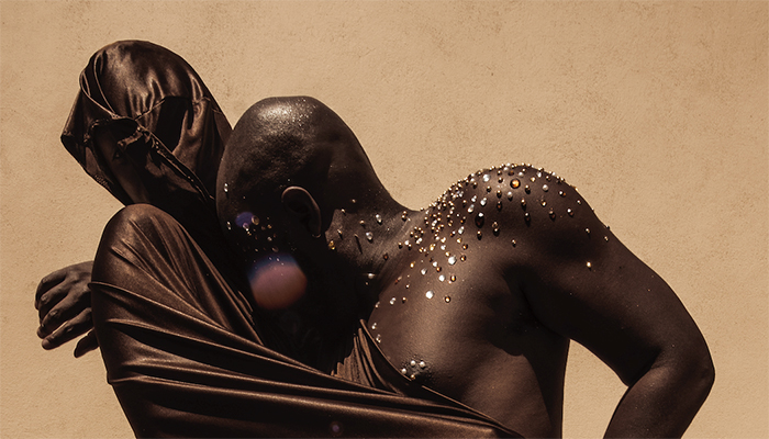 Nile Harris (left) and jaamil olawale kosoko (right) embracing one another. Harris’s face and body are covered with shimmery brown fabric. kosoko—who has dark brown skin and silver, gold, and brown gems on their bare shoulder, neck, and chest—is leaning their bare head on Harris’s chest.