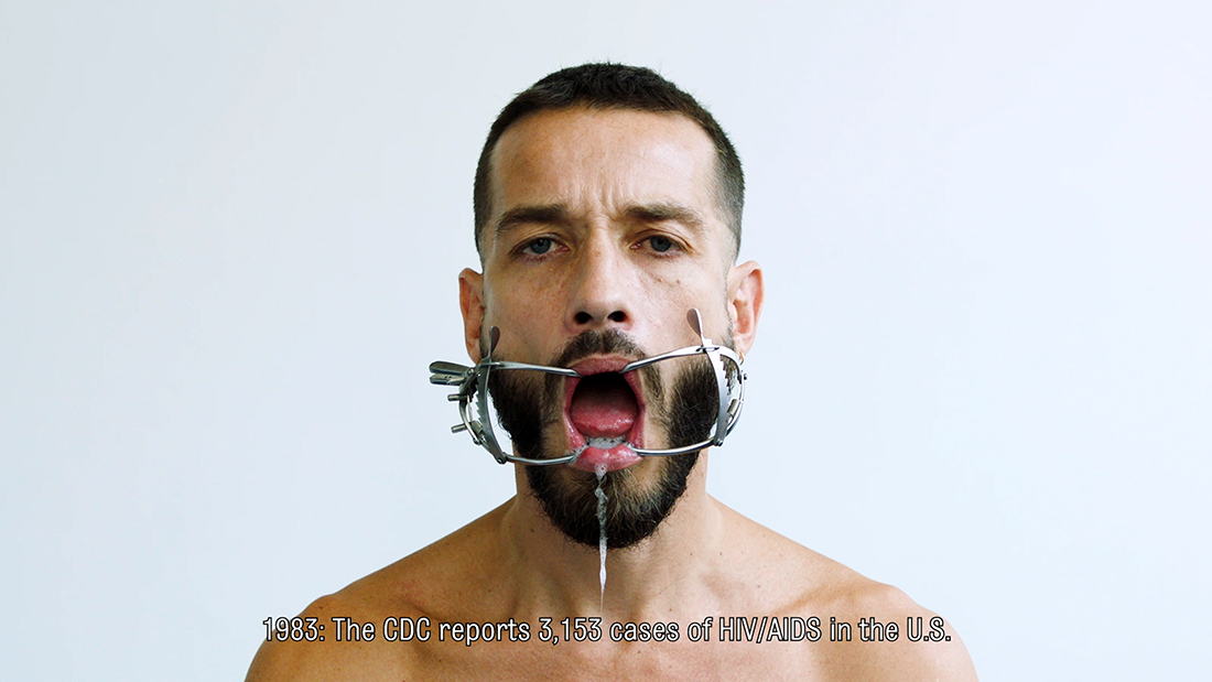 Carlos Motta from his shoulders up, with a dental gag in his mouth and the following text across his bare chest: "1983: The CDC reports 3,153 cases of HIV/AIDS in the U.S."