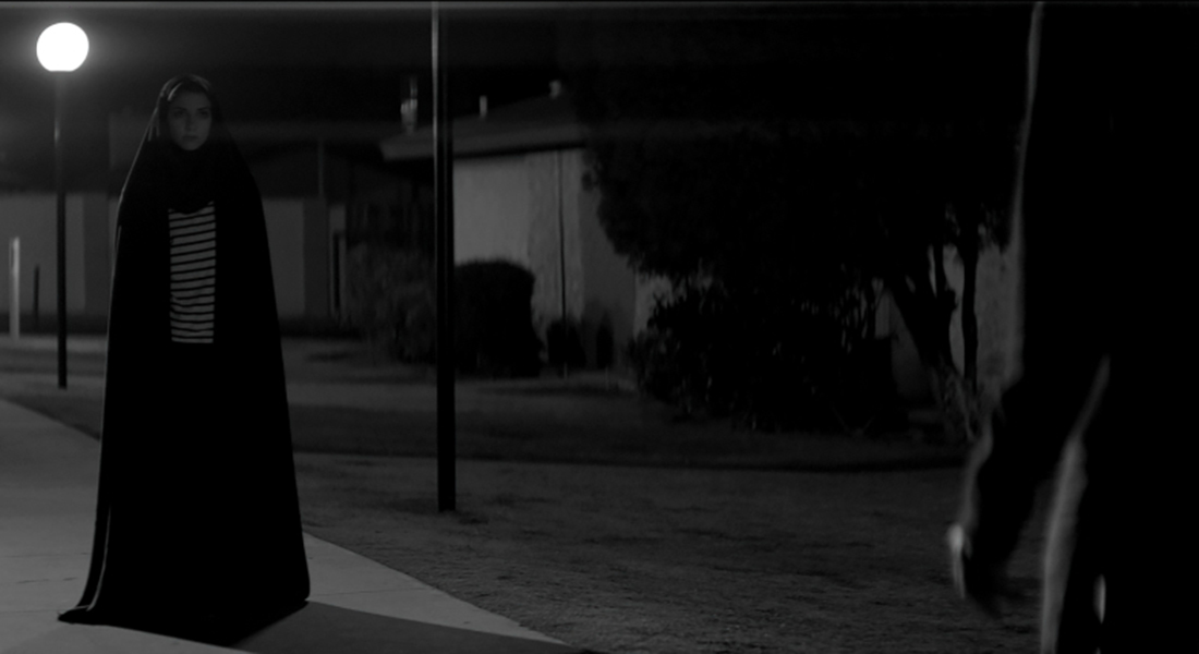 The Girl standing on a sidewalk with a streetlight shining behind her. She is wearing a long, dark chador and a striped top. The figure of a person is visible on the right side of the image, and the Girl is looking straight at them from the left.