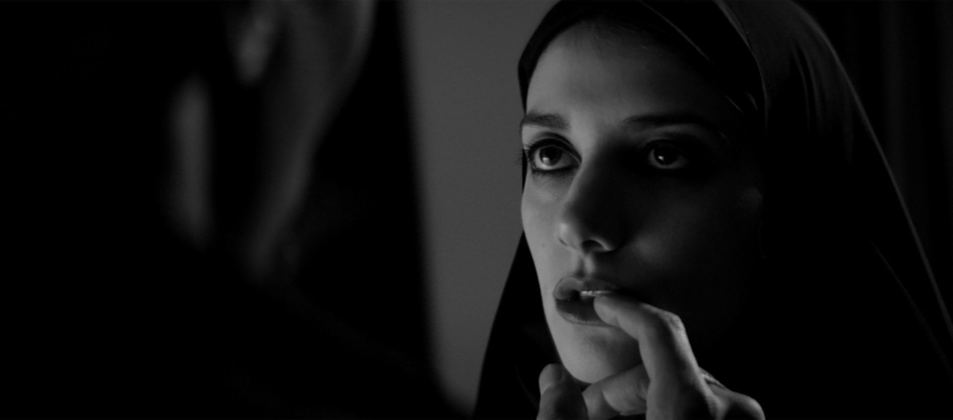 The Girl looking into the distance and wearing a dark chador. The left side of the image is blacked out due to someone is standing in front of the Girl. The unknown person’s finger is touching the Girl’s bottom lip.