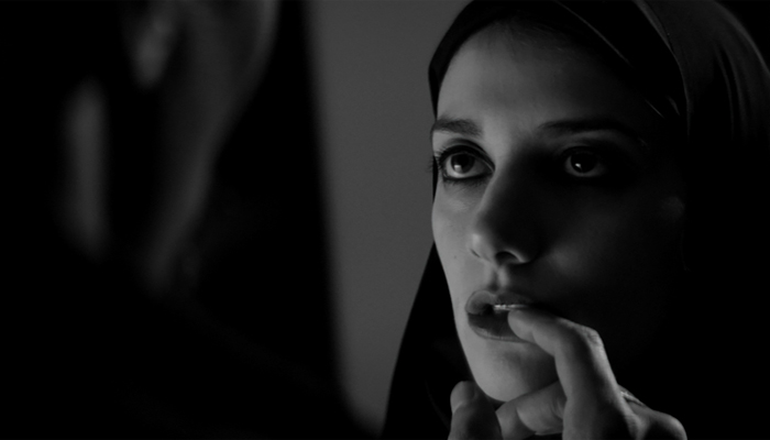 The Girl, who is wearing a dark chador. She is looking ahead at someone barely visible in front of her. The unknown person’s finger is touching the Girl’s bottom lip.
