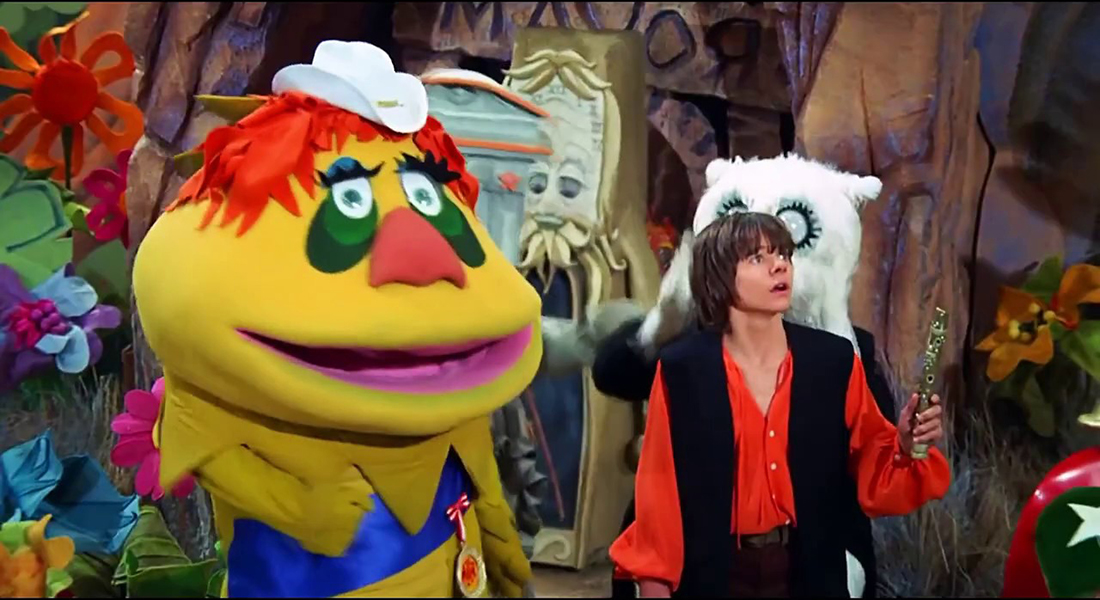 HR Pufnstuf, a puppet in bright bold colors and a very large head including green eyes and tiny white hat stands next to a young boy in a red shirt and black vest holding a flute