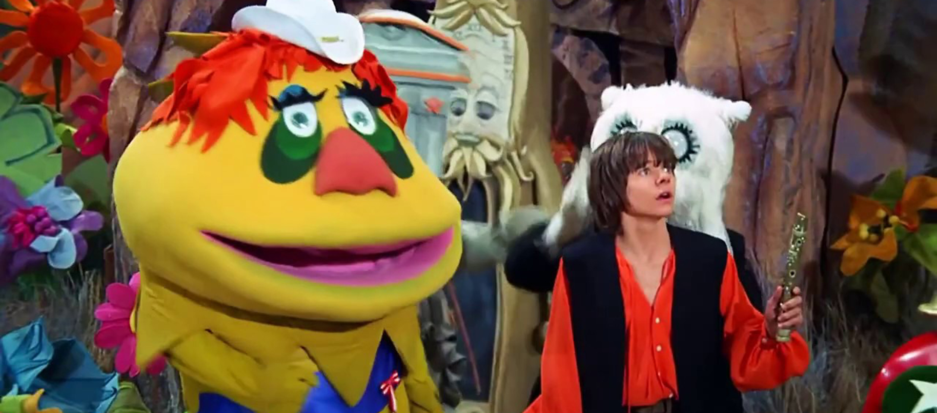 HR Pufnstuf, a puppet in bright bold colors and a very large head including green eyes and tiny white hat stands next to a young boy in a red shirt and black vest holding a flute