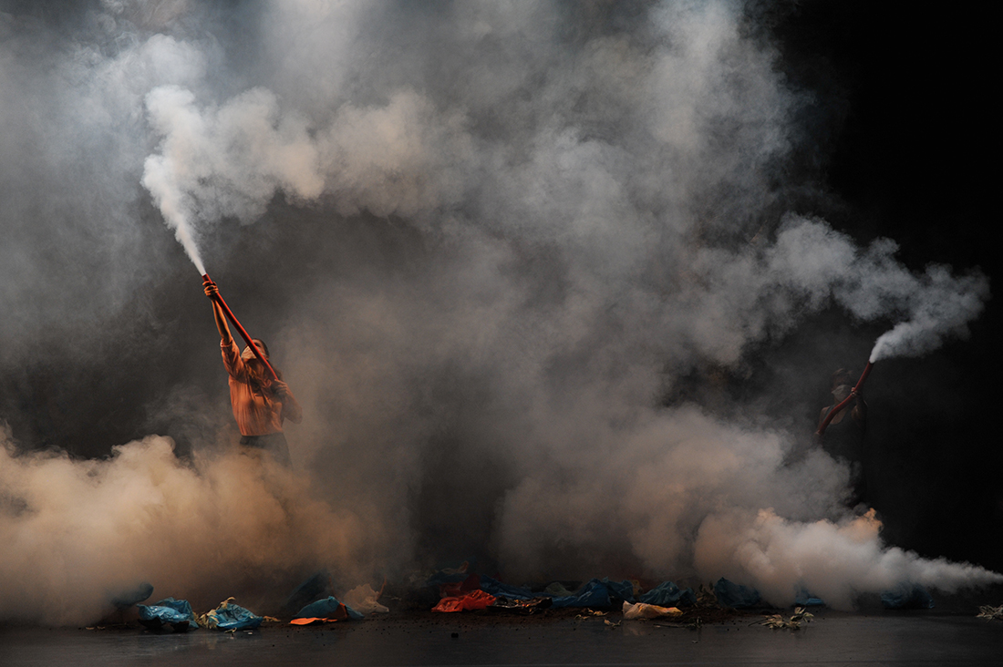 Two performers point red hoses into the air, emitting white smoke across the stage.