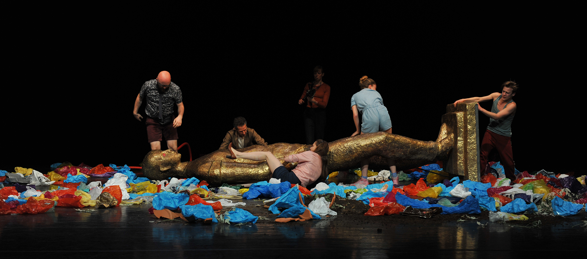 Six performers in various outfits surround a large, toppled gold statue of a deity lying on a stage that is covered in colorful plastic bags.