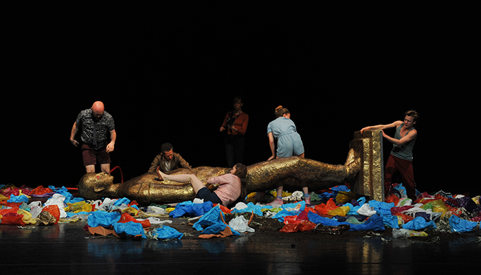 Six performers in various outfits surround a large, toppled gold statue of a deity lying on a stage that is covered in colorful plastic bags.