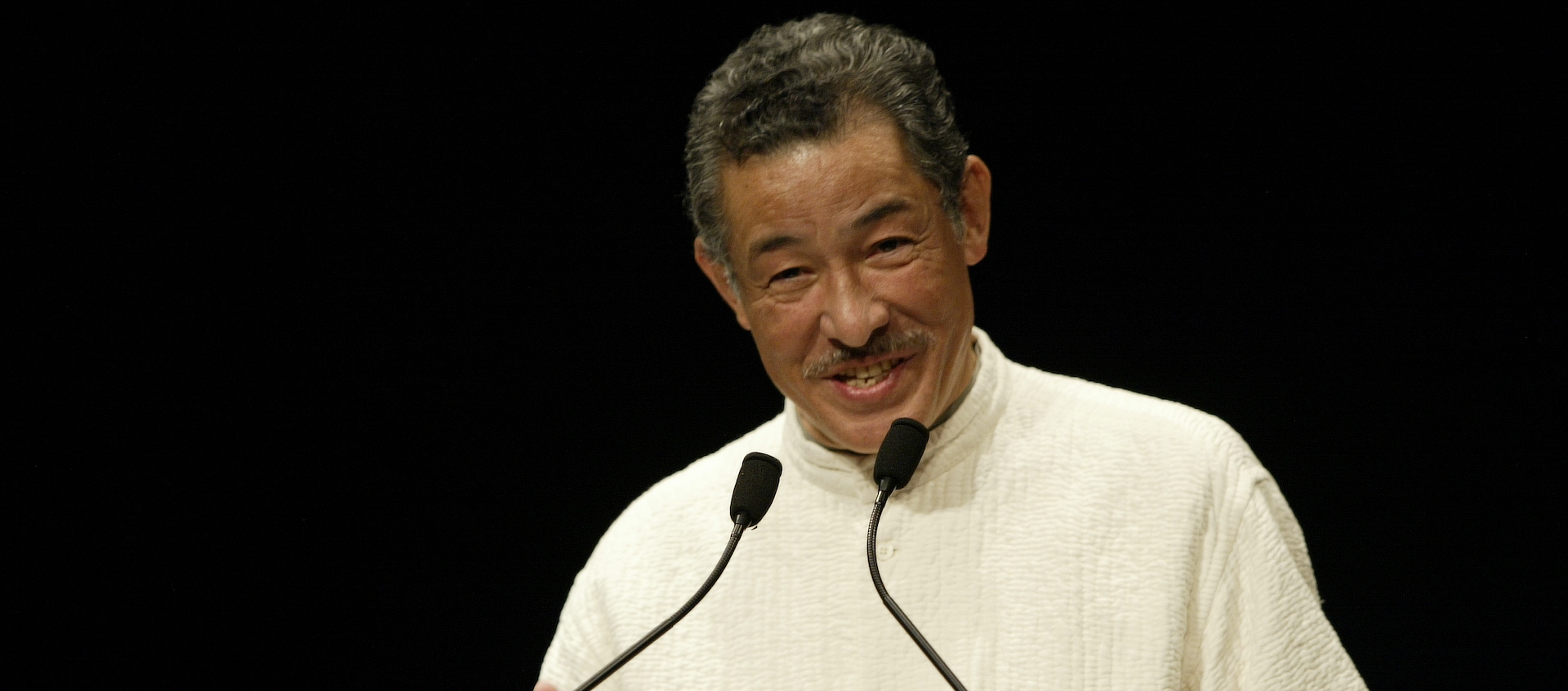 Japanese fashion designer Issey Miyake speaks into two microphones extending up from an unseen podium. He's wearing an off-white turtleneck and is standing in front of a black background