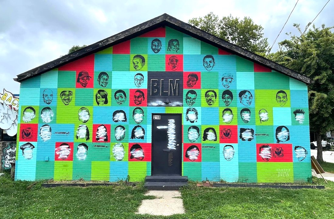 The west-facing exterior wall of The Vanderelli Room, which is covered with multicolored squares featuring black stencils of faces of Black victims of police violence with the letters "BLM" in a black square above a door at the center. More than 20 of the faces are covered by white spray paint.