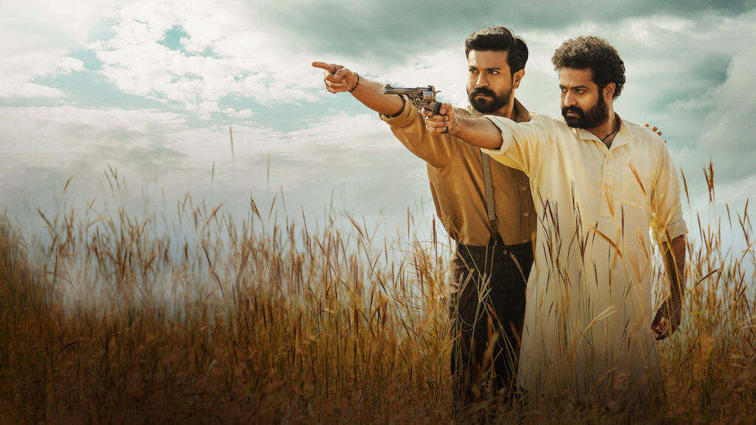 Two bearded Indian men stand beside each other in a field of wheat up to their waists. Both are looking and pointing to something unseen to their right; the man in the foreground is holding a gun