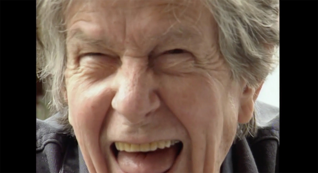 The face of a person with white, shaggy hair just over their ears. They are laughing and their face is scrunched up with their eyes narrowed into slits and their mouth agape.