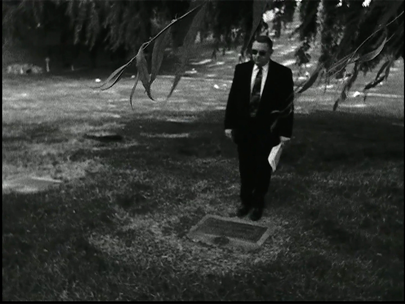 Black-and-white photo of a person, who is wearing a dark suit and glasses, standing in front of a grave. Tree branches appear in the foreground.
