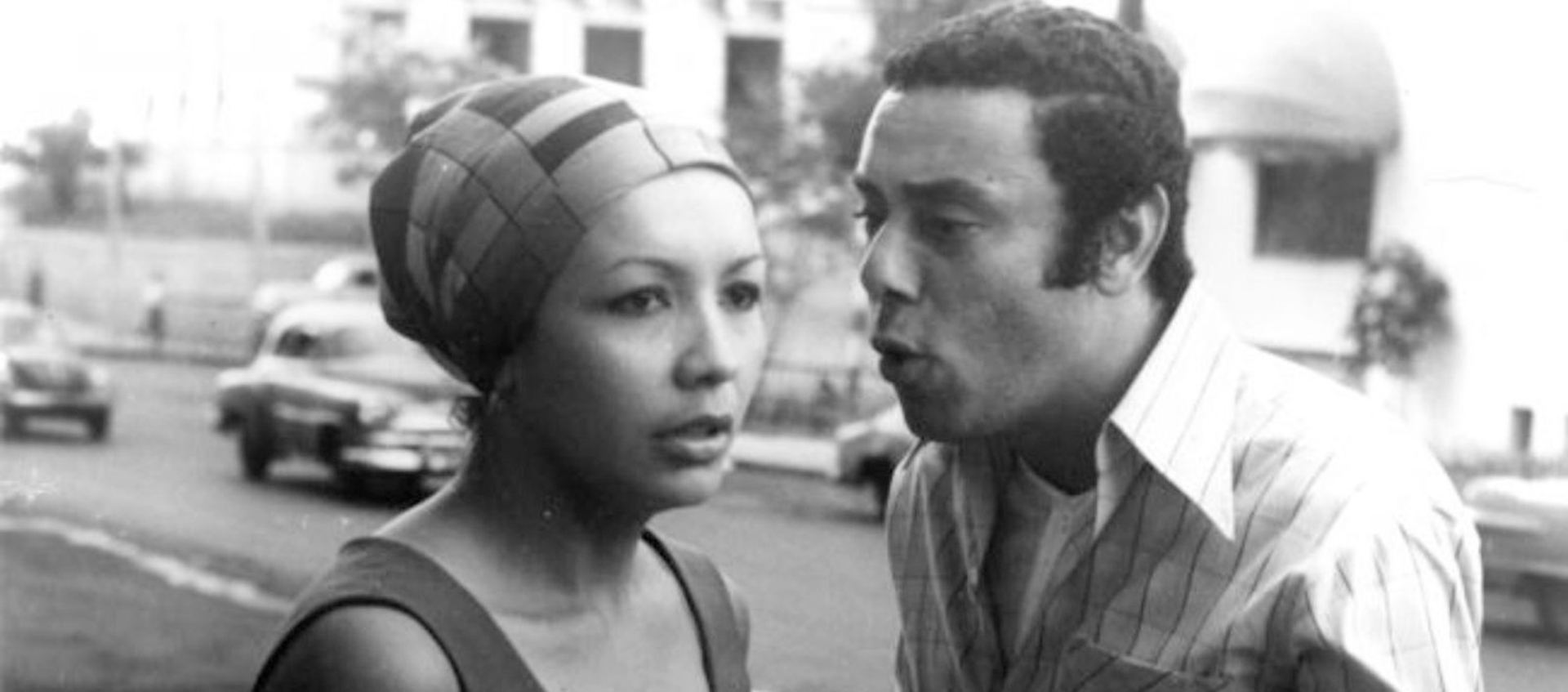 A black-and-white image of a woman and man on the street. The woman wears a cap covering her head and hair and wears a tank top. To her right a man in a button down shirt and t-shirt underneath speaks to her.