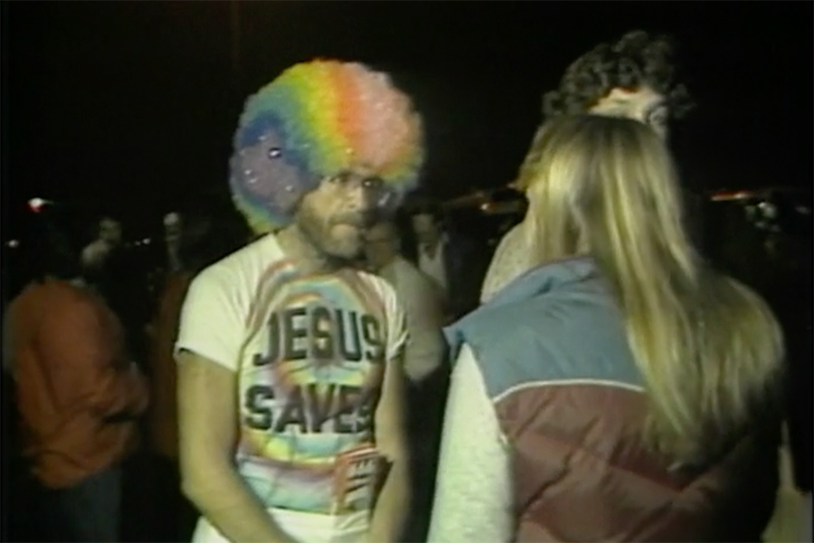 A man with a brightly colored rainbow wig wears a colorful t-shirt with the words "Jesus Saves" on the front. He has a short beard and is speaking with a person with long hair and a winter vest on.
