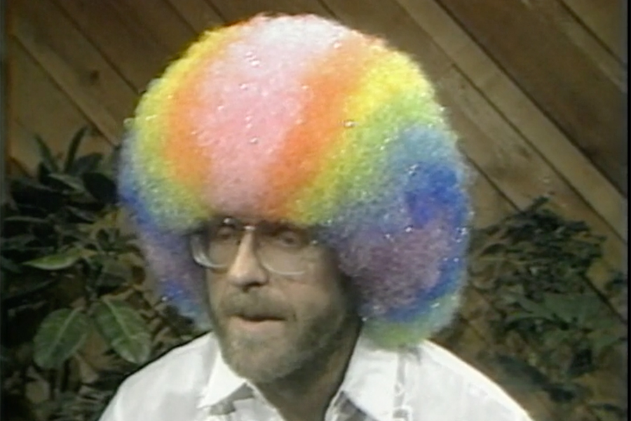 A man with a brightly colored rainbow wig is seen from the shoulders up. He wears eyeglasses and sits in front of a brown, wood-paneled wall.