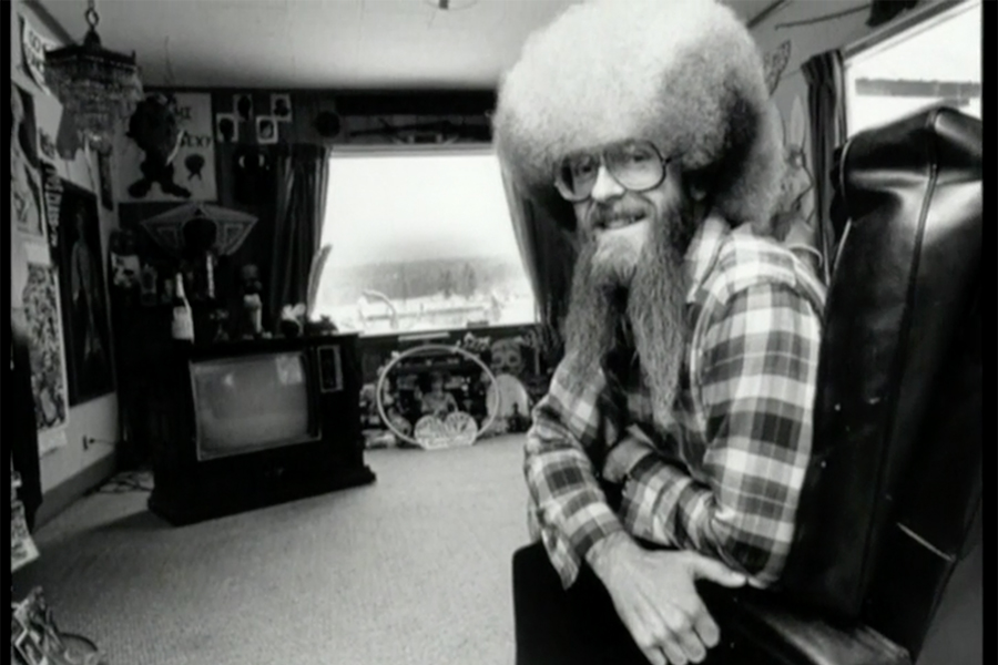 A man in a rainbow wig wears a plaid shirt. He is seated in a room in front of a television.