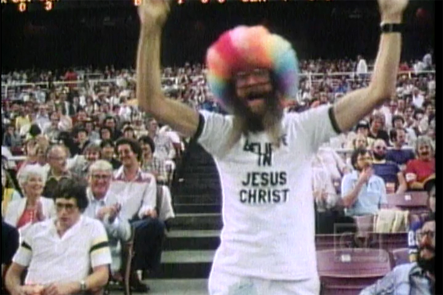 A man in a rainbow wig stands among a crowd of spectators at a sporting event. He wears a white t-shirt with the words "Believe in Jesus Christ" on it. He is standing and his arms are outstretched and pointed upward.