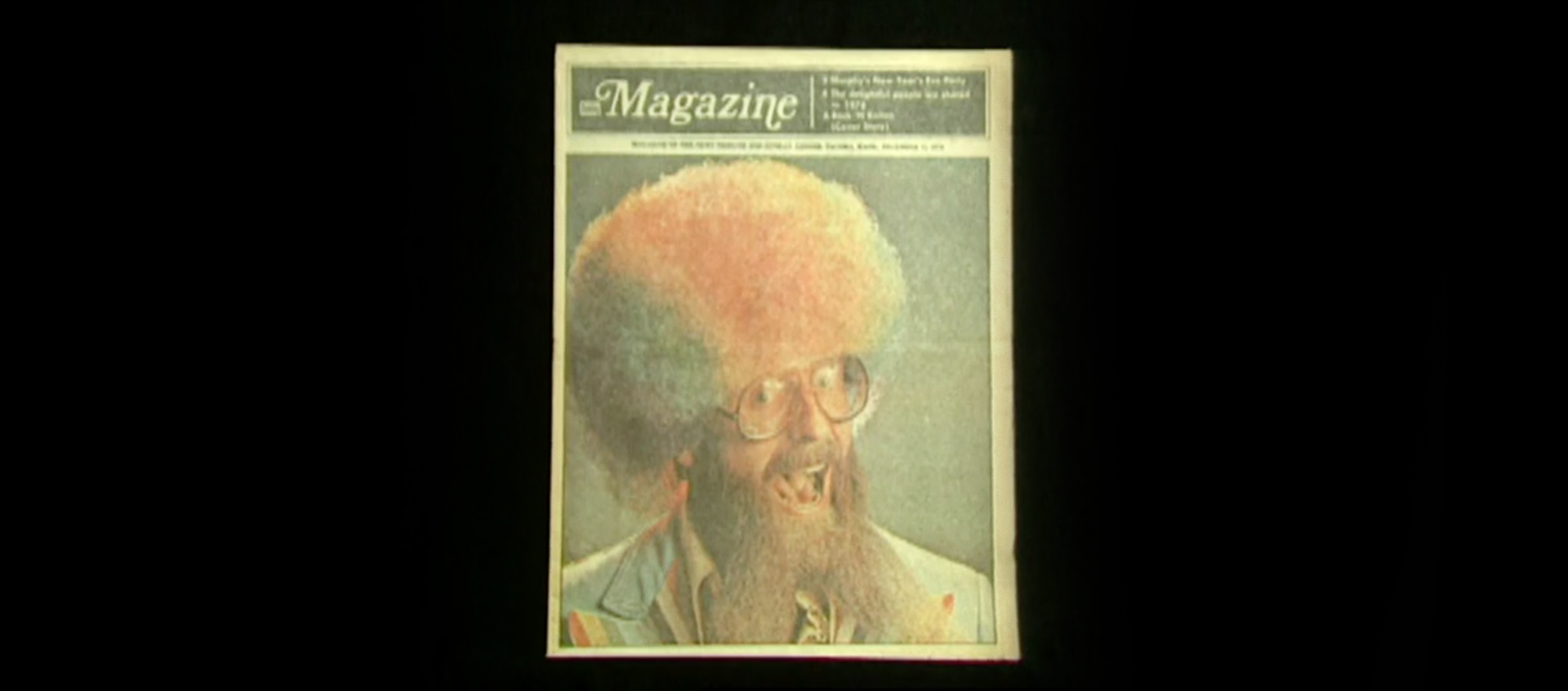 A magazine called "Magazine" depicts a smiling man, mouth open, in a brightly colored rainbow wig. He wears eyeglasses and has a beard that goes down to his chest.