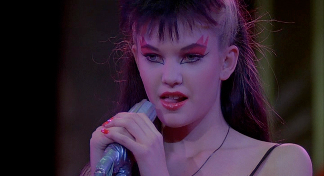 A punk singer with their head shaved on the sides, but long in the back wears bright red eye shadow that swoops upward over their brows. They grip a tape covered microphone with their hands that have pink nail polish on each finger. They have bright red lips and their tongue is licking the lower left side of the mouth.