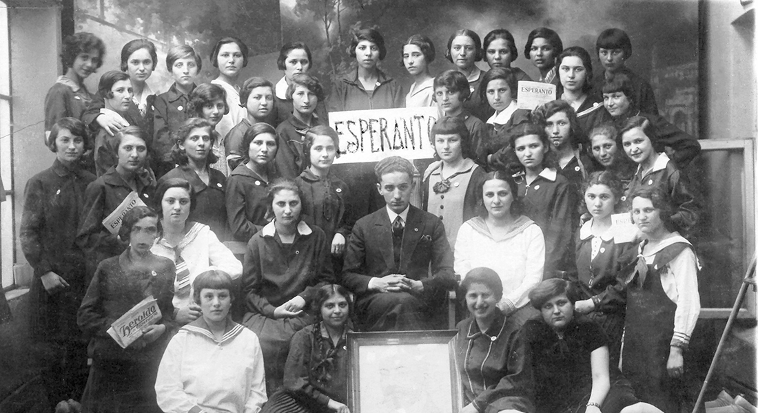 A black-and-white image of a large group of young people facing toward the camera. In the center of this group shot, a person holds a handwritten sign that says "Esperanto."