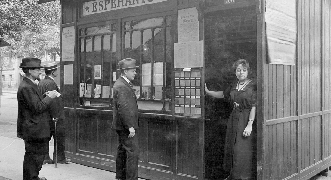 A woman in a dark dress stands in the doorway of a kiosk on the street. Three men, all in hats, stand in line waiting to speak with her.