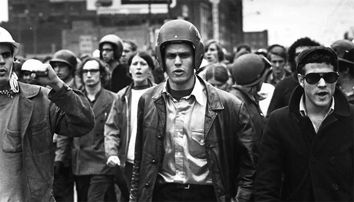 Black-and-white photo of a crowd marching. The person in the front middle is wearing a helmet and leather jacket and has a furrowed brow.