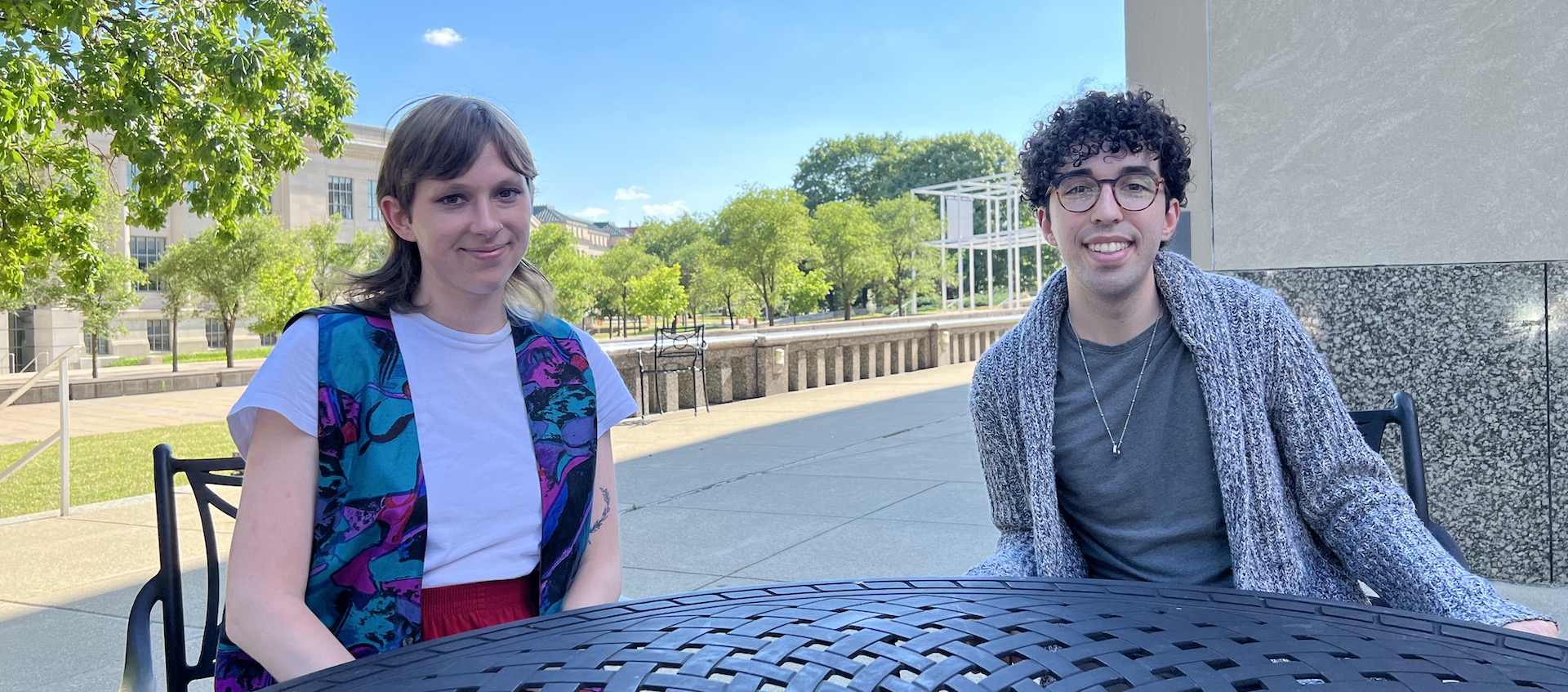 Laurel Hilliard and Austin Dunn sit at a table outside Mershon Auditorium with the Wexner Center in the background. Laurel has shoulder length light brown hair and is wearing a white top with a dark vest. Austin has curly dark hair and glasses. They are wearing a collared shirt and a cardigan sweater.