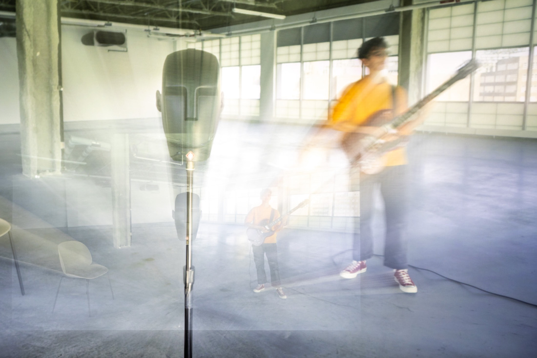 Still from the live documentary 32 Sounds. A head-shaped microphone on a stand is in the foreground of a mostly empty, widow-lined space. Musician JD Samson stands alone, playing an electric guitar. She is wearing a yellow top, gray paints, and red sneakers. The image is refracted with light, creating a smaller image of Samson to the left of where she stands. 