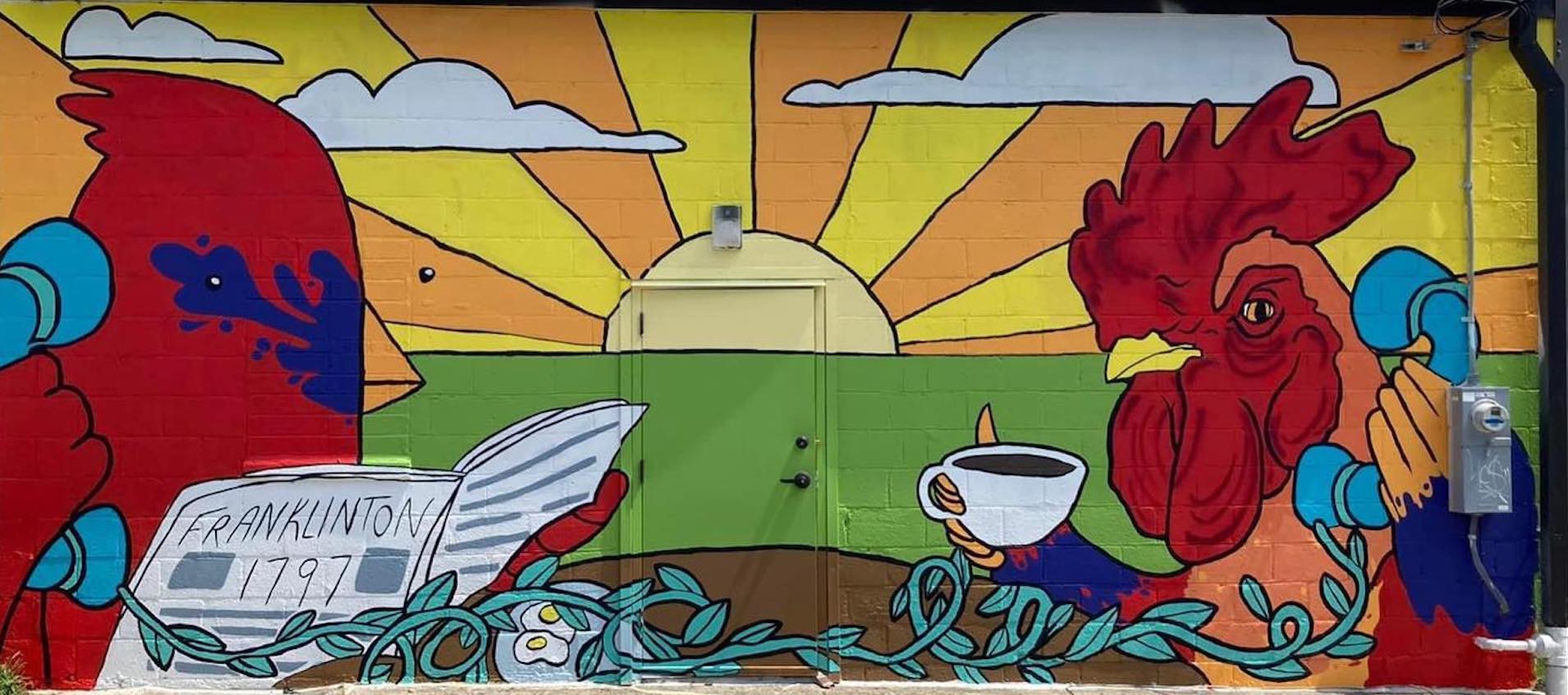 An exterior building mural, featuring a cardinal on the left reading a newspaper and holding a blue, classic telephone receiver to its ear. To its right, a rooster sits holds a cup of coffee and an identical phone receiver. Between them is a brown table and a leafy green vine connecting the phone receivers. A green field and a a rising sun with yellow and orange beams extending from it can be seen behind and between the two birds.