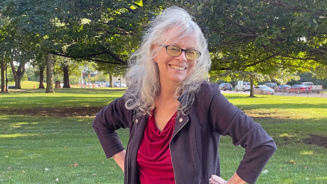 Candice Watkins stands in a grassy park with trees in the background. She has long white hair and dark-rimmed glasses. She's wearing a red top and black jacket and is smiling, with her hands placed on her hips.