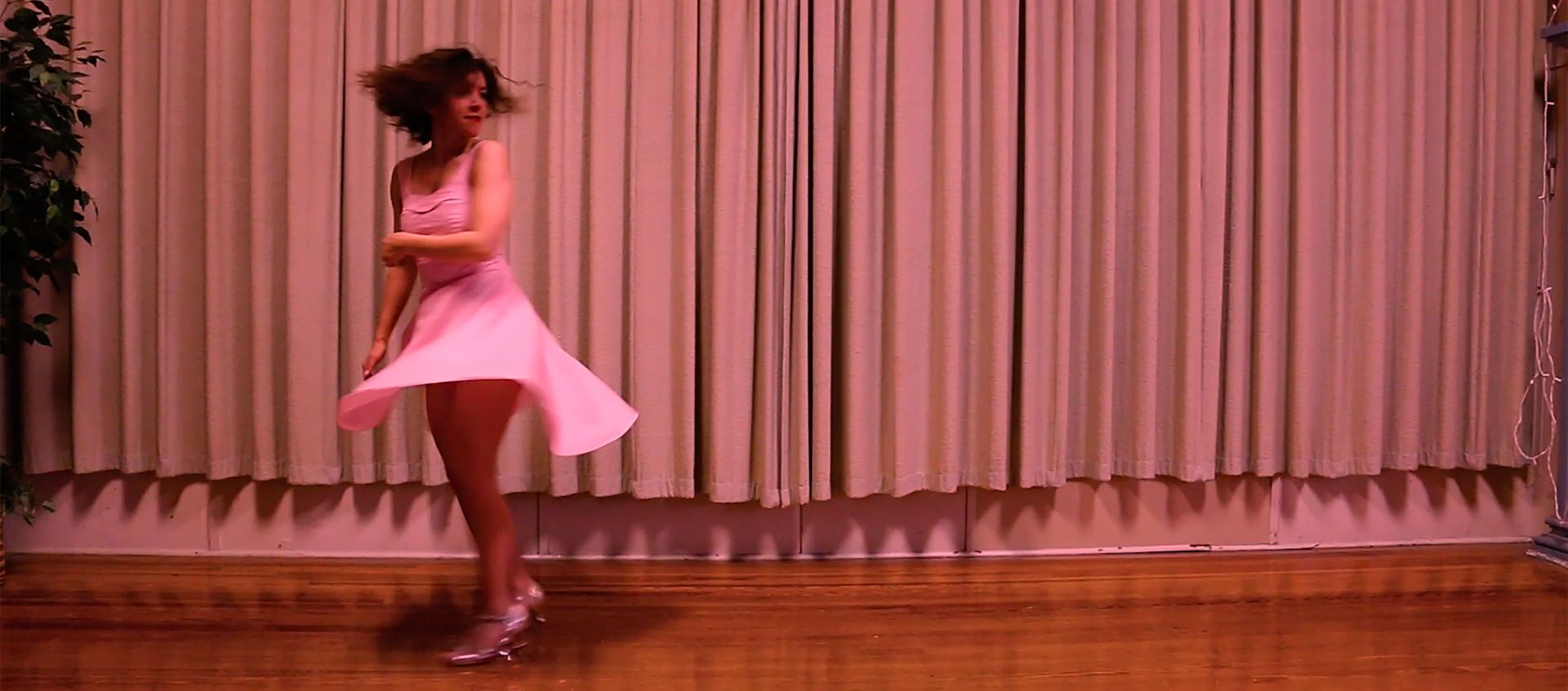 Still from Am Schmidt's remake of Dirty Dancing. Schmidt stands alone on a stage with a curtain backdrop. She is left of center, wearing a pink dress and shiny heeled shoes and she's captured mid-twirl.