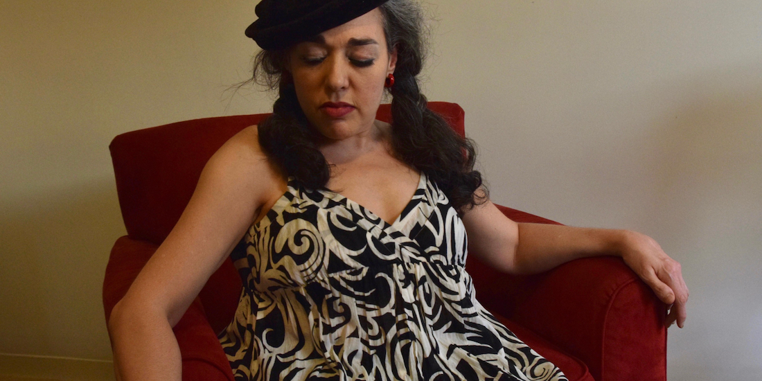 Photo of singer-songwriter and activist Tracy Marie. She is sitting on an orange velvet chair. She has long dark hair. She wears a sleeveless v-neck dress with a swirling black and white pattern. She is looking down.