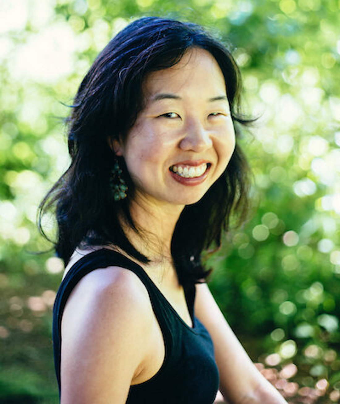 Photo of writer Shelley Wong. She's seen from the shoulders up, her body facing to the right while her face is turned toward the camera, smiling. She has shoulder-length dark hair and is wearing a black tank top. Leafy trees are seen in the background.