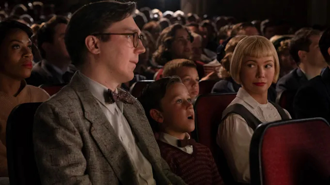 A still from the film The Fabelmans with Paul Dano in a bowtie and glasses seated next to a small child and Michelle Williams