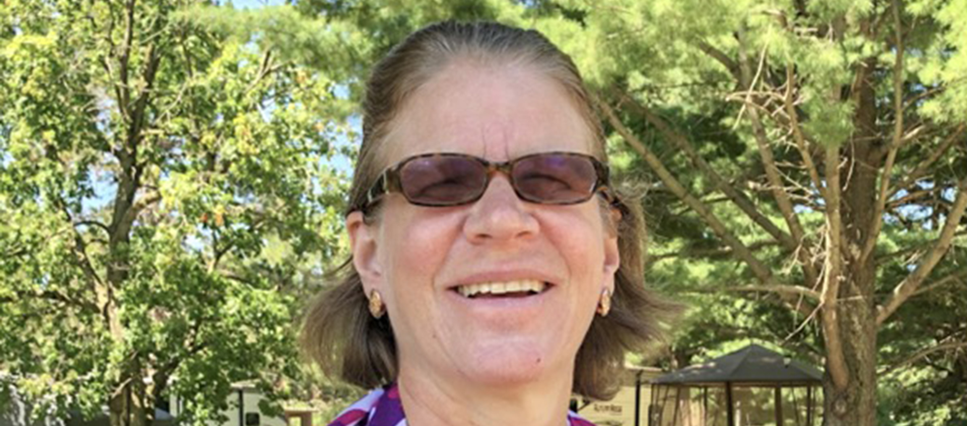 Smiling woman with white skin and chin-length wavy blond hair pulled back on top. She wears dark glasses and a colorful scarf over her shoulders and is standing in front of verdant trees spanning a light blue sky.