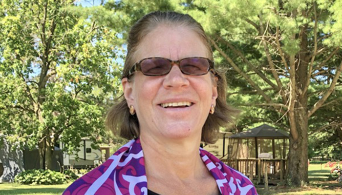 Smiling woman with white skin and chin-length wavy blond hair pulled back on top. She wears dark glasses and a colorful scarf over her shoulders and is standing in front of verdant trees spanning a light blue sky.