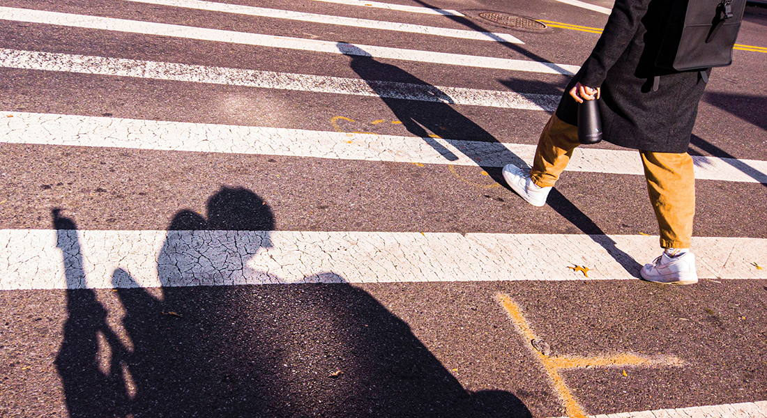 Point-of-view shot featuring the shadow of a person in a wheelchair seen on a crosswalk. To the right of the shadow is the lower half of a person who is walking. 