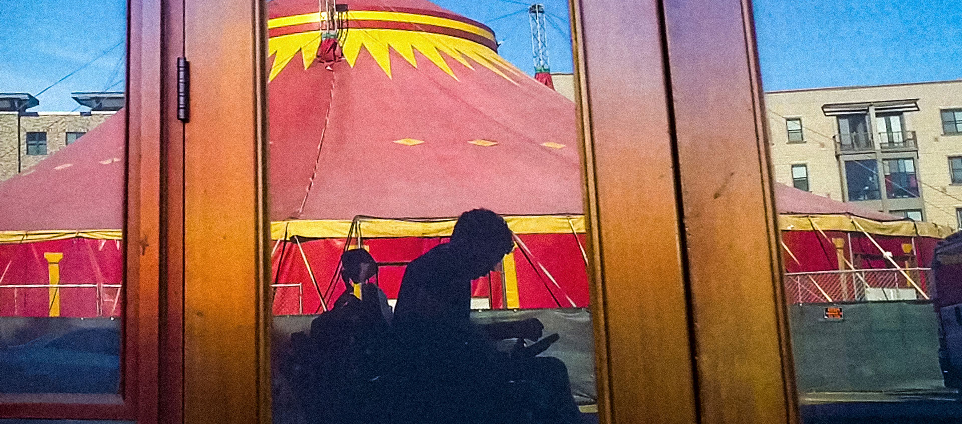 In a reflection of an unmarked storefront is a grayish silhouette of a man using an electric wheelchair. Behind the man is a spectacular red-and-yellow circus tent.