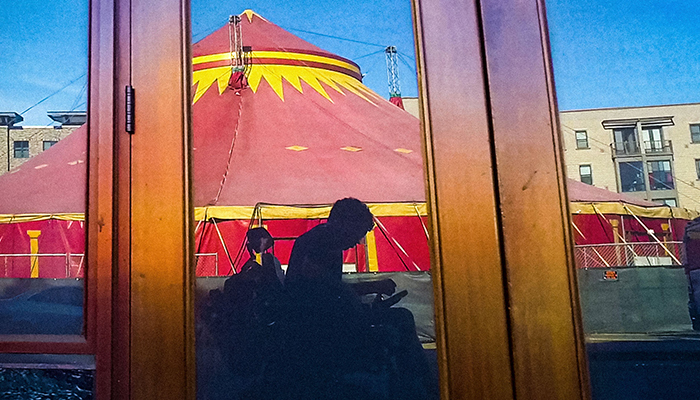In a reflection of an unmarked storefront is a grayish silhouette of a man using an electric wheelchair. Behind the man is a spectacular red-and-yellow circus tent.