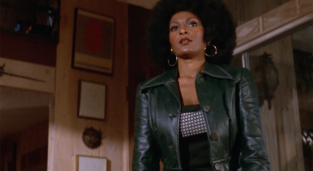 Pam Grier from the waist up in a room with wooden walls with art hanging from them. She wears a leather jacket and has her hair styled in an afro.