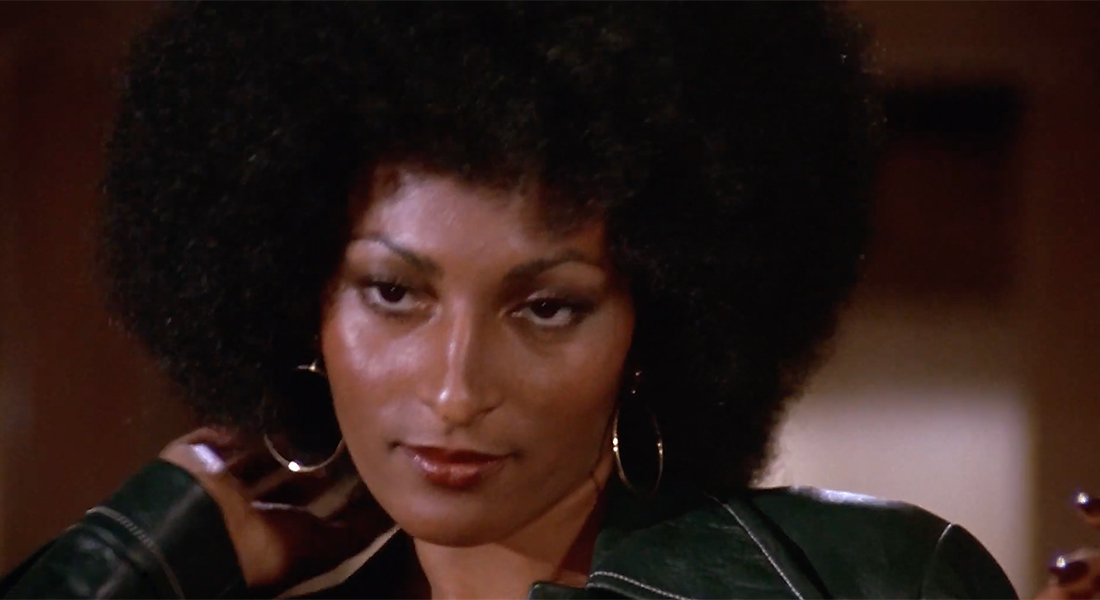 Close-up of Pam Grier, whose right hand is touching her hair, which is styled in an afro. She wears silver hoop earrings and reddish lipstick.